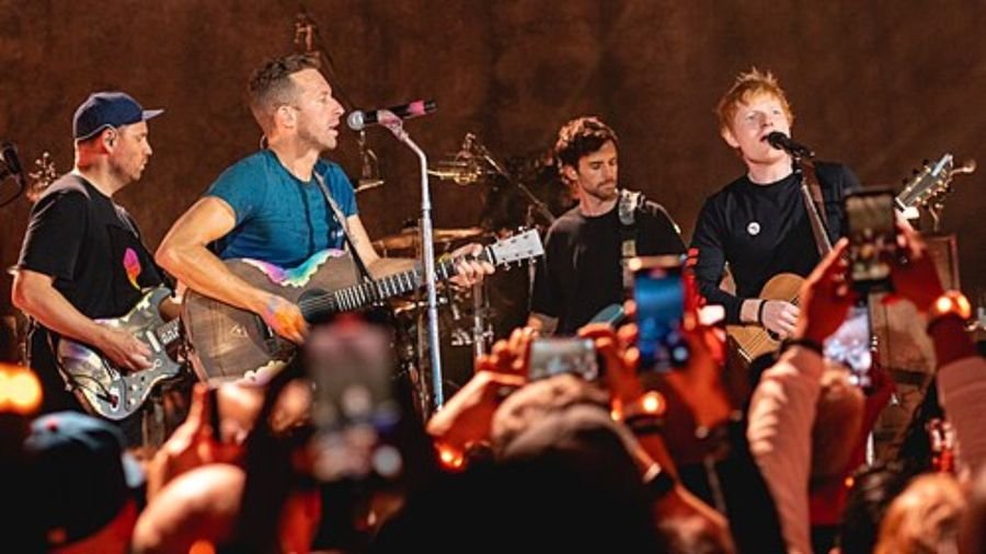 Four musicians performing on stage in front of a crowd holding up phones, shown in the Multiply album section